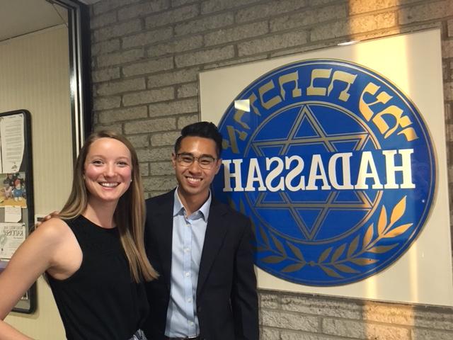 Two students who traveled to Hadassah stand, smiling, in front of a large plaque of the Hadassah Medical Center logo.